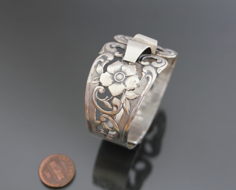 May include: Silver bracelet with a floral design. The bracelet is open and has a clasp on the side. The design features a large flower with a smaller flower on the side.