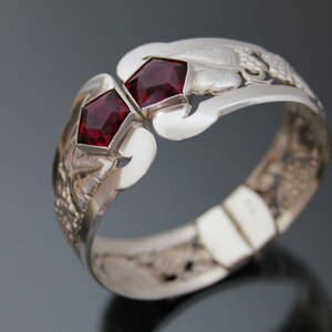 Puede incluir: Un brazalete de plata con un dise&ntilde;o intrincado que presenta dos piedras preciosas rojas. El brazalete est&aacute; abierto y tiene un cierre con bisagra.