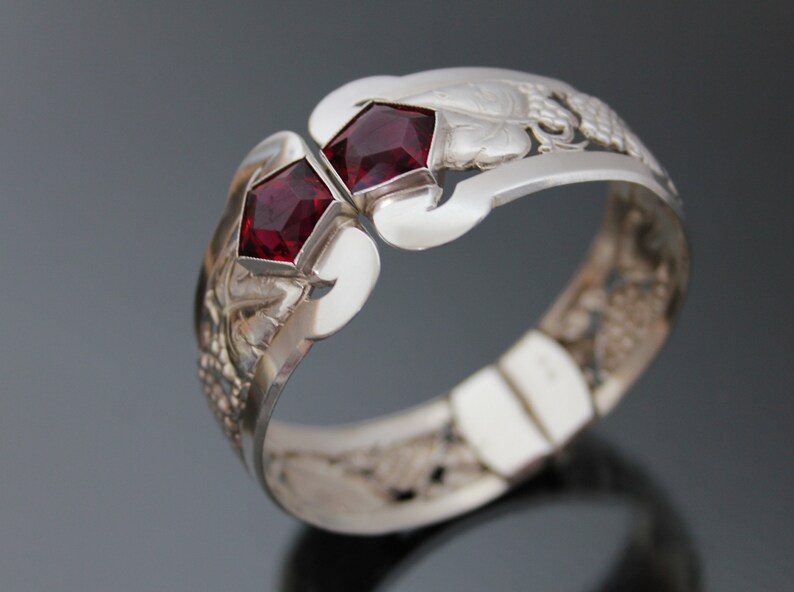 Puede incluir: Un brazalete de plata con un dise&ntilde;o intrincado que presenta dos piedras preciosas rojas. El brazalete est&aacute; abierto y tiene un cierre con bisagra.