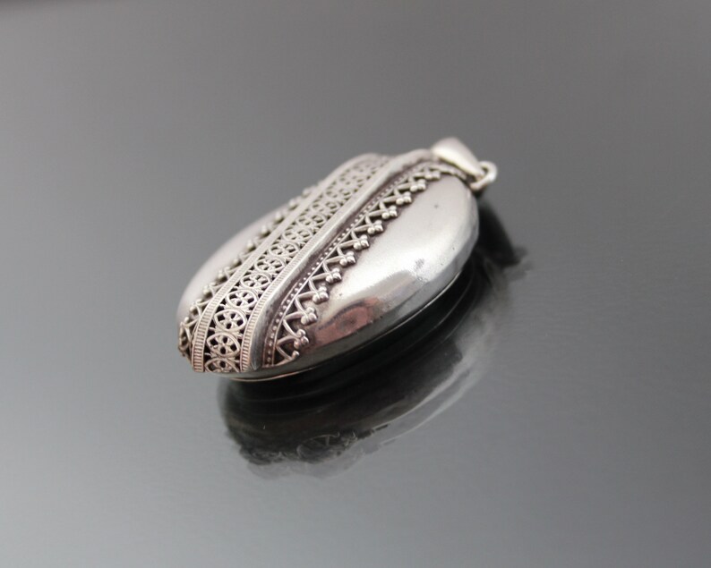 K&ouml;nnte beinhalten: Ein silbernes Medaillon mit einem komplizierten Design am Rand. Das Medaillon ist oval geformt und hat eine glatte Oberfl&auml;che.