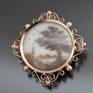 May include: A gold-toned brooch with a delicate, ornate frame. The frame encloses a round glass cameo with a hand-painted scene of a church and a tree. The scene is created using hair, a technique known as hair work.