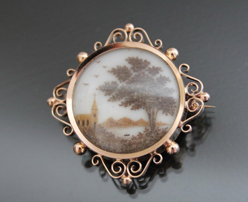 May include: A gold-toned brooch with a delicate, ornate frame. The frame encloses a round glass cameo with a hand-painted scene of a church and a tree. The scene is created using hair, a technique known as hair work.