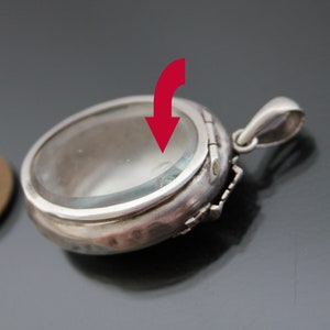 K&ouml;nnte beinhalten: Silberner Anh&auml;nger mit einem klaren Glasdeckel und einem Scharnier auf der R&uuml;ckseite. Der Anh&auml;nger ist oval geformt und hat eine zarte Blumenverzierung am Rand.