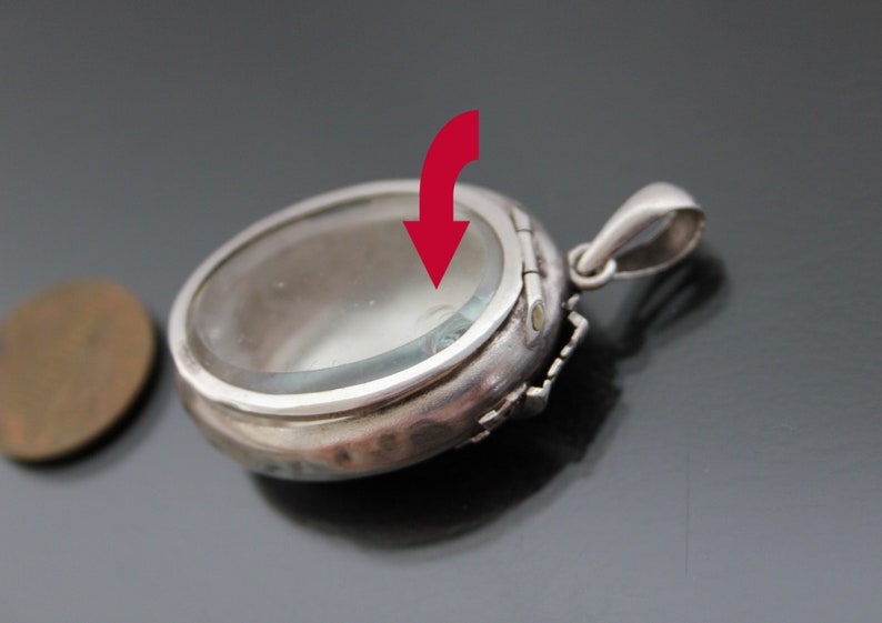 K&ouml;nnte beinhalten: Silberner Anh&auml;nger mit einem klaren Glasdeckel und einem Scharnier auf der R&uuml;ckseite. Der Anh&auml;nger ist oval geformt und hat eine zarte Blumenverzierung am Rand.