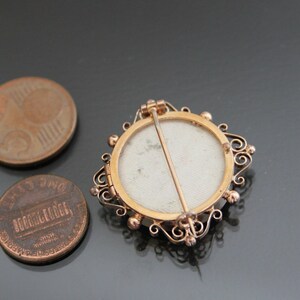 May include: A gold-toned locket with a white cameo and a filigree design. The locket is open and the back is visible. The locket is on a gray surface.