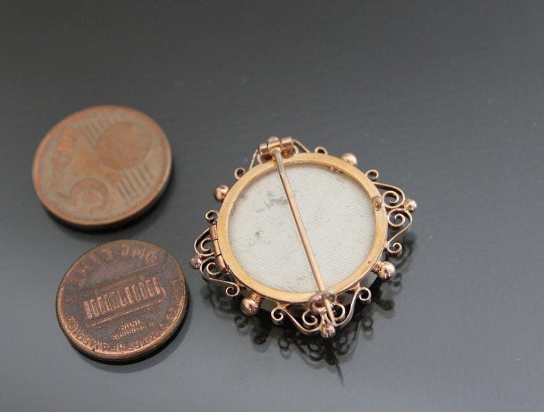 May include: A gold-toned locket with a white cameo and a filigree design. The locket is open and the back is visible. The locket is on a gray surface.