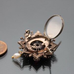 Op de afbeelding: Een zilveren en gouden broche met een stervormig ontwerp en een klein parelhangertje. De broche heeft een scharnierend deksel dat opengaat om een kleine spiegel te onthullen.