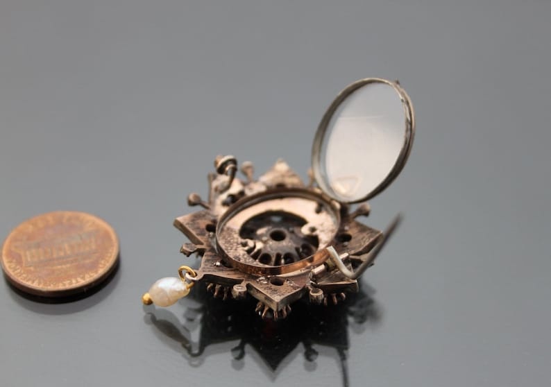 Op de afbeelding: Een zilveren en gouden broche met een stervormig ontwerp en een klein parelhangertje. De broche heeft een scharnierend deksel dat opengaat om een kleine spiegel te onthullen.