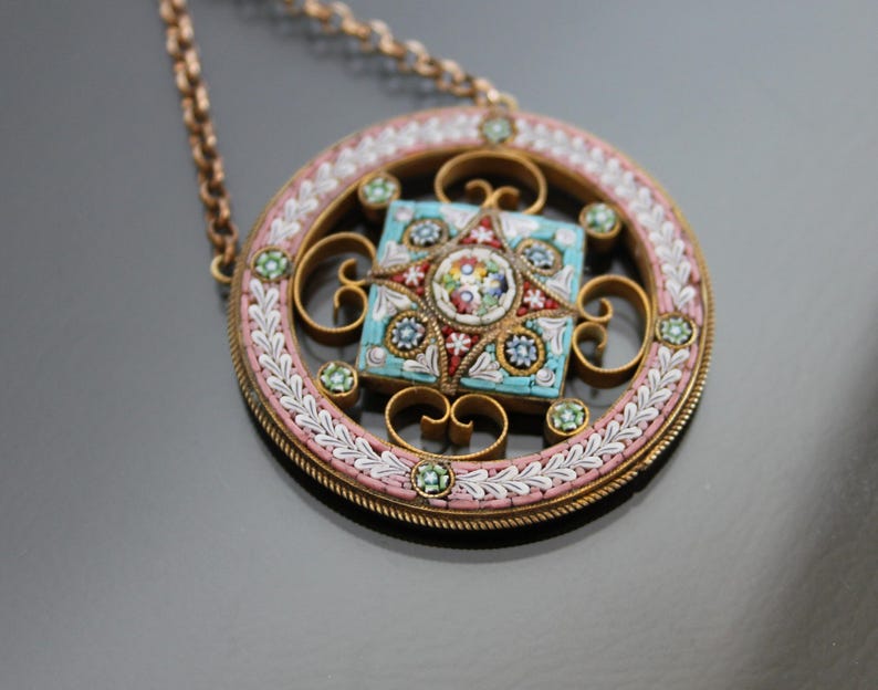 May include: A round pendant necklace with a delicate gold chain. The pendant features a square mosaic design with a star in the center. The mosaic is made of small, colorful tiles in shades of blue, green, pink, and red.