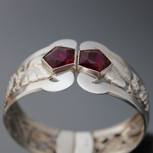 Puede incluir: Un brazalete de plata con un dise&ntilde;o intrincado que presenta dos piedras preciosas rojas. El brazalete est&aacute; abierto y tiene un aspecto delicado y ornamentado.