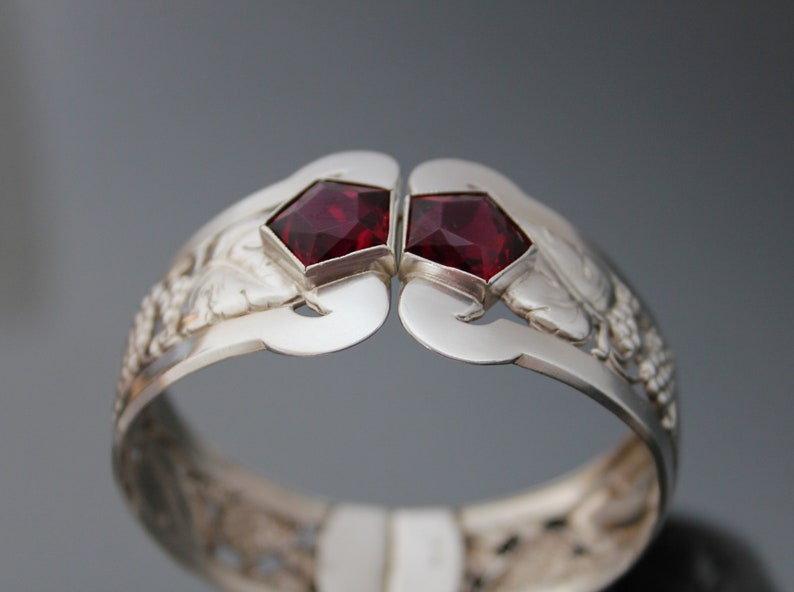 Puede incluir: Un brazalete de plata con un dise&ntilde;o intrincado que presenta dos piedras preciosas rojas. El brazalete est&aacute; abierto y tiene un aspecto delicado y ornamentado.