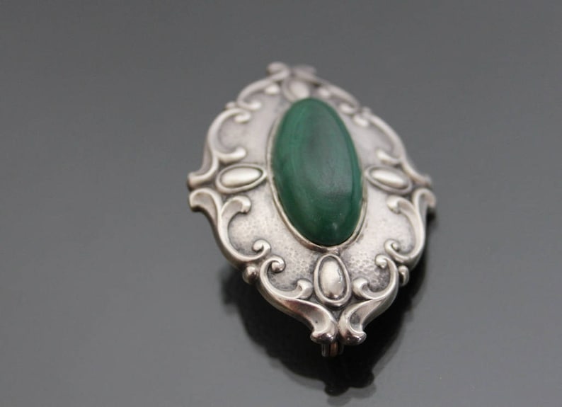 May include: A silver brooch with an oval-shaped green malachite stone set in a detailed, ornate design.