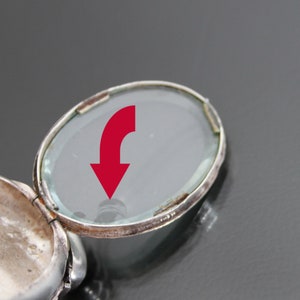 K&ouml;nnte beinhalten: Ein silbernes Medaillon mit einem Scharnierdeckel und einem transparenten Glaseinsatz. Das Medaillon ist ge&ouml;ffnet und zeigt das Innere des Medaillons.