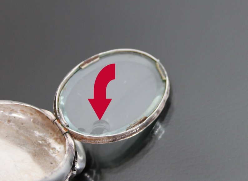K&ouml;nnte beinhalten: Ein silbernes Medaillon mit einem Scharnierdeckel und einem transparenten Glaseinsatz. Das Medaillon ist ge&ouml;ffnet und zeigt das Innere des Medaillons.