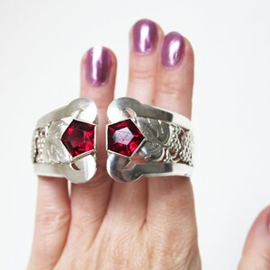 Puede incluir: Un brazalete de plata con dos acentos de piedras preciosas rojas. El brazalete est&aacute; abierto y tiene un dise&ntilde;o floral.