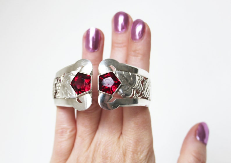 Puede incluir: Un brazalete de plata con dos acentos de piedras preciosas rojas. El brazalete est&aacute; abierto y tiene un dise&ntilde;o floral.
