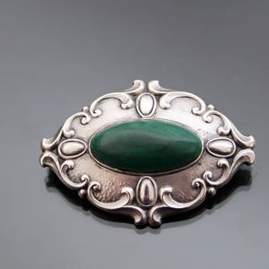 May include: A silver brooch with an oval-shaped green malachite stone set in a detailed, ornate design.
