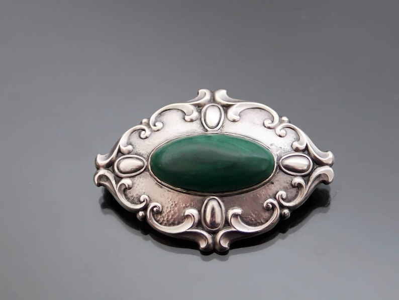 May include: A silver brooch with an oval-shaped green malachite stone set in a detailed, ornate design.