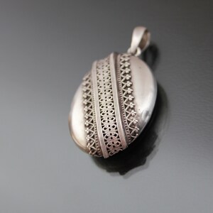 K&ouml;nnte beinhalten: Ein silbernes Medaillon mit einem komplizierten, verzierten Design. Das Medaillon ist oval geformt und hat oben eine Schlaufe zum Anbringen einer Kette.