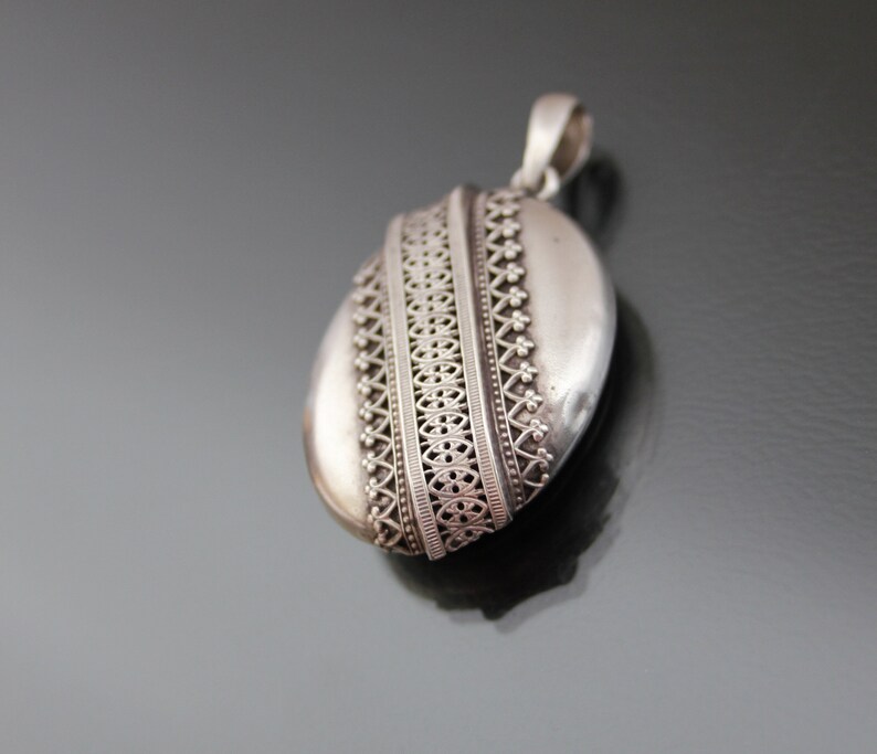 K&ouml;nnte beinhalten: Ein silbernes Medaillon mit einem komplizierten, verzierten Design. Das Medaillon ist oval geformt und hat oben eine Schlaufe zum Anbringen einer Kette.