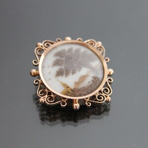 May include: A gold-toned brooch with a delicate filigree design. The center of the brooch features a round, white cameo with a brown and white image of a tree and a bird.