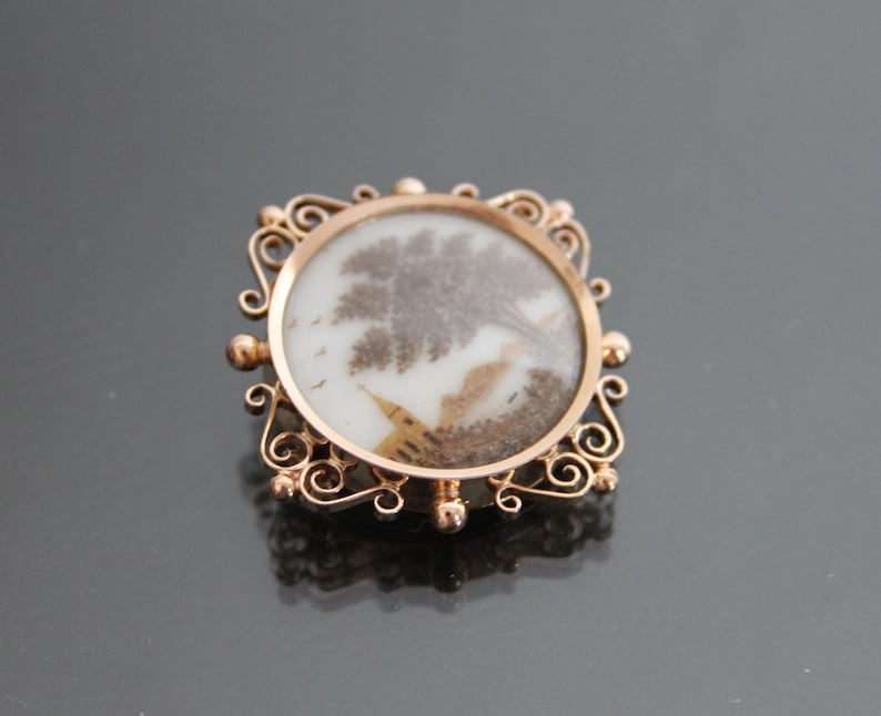 May include: A gold-toned brooch with a delicate filigree design. The center of the brooch features a round, white cameo with a brown and white image of a tree and a bird.