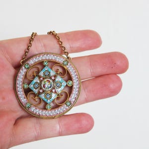 May include: A round pendant necklace with a delicate gold chain. The pendant is made of white, pink, and blue mosaic tiles arranged in a geometric pattern. The pendant is surrounded by a gold filigree design.