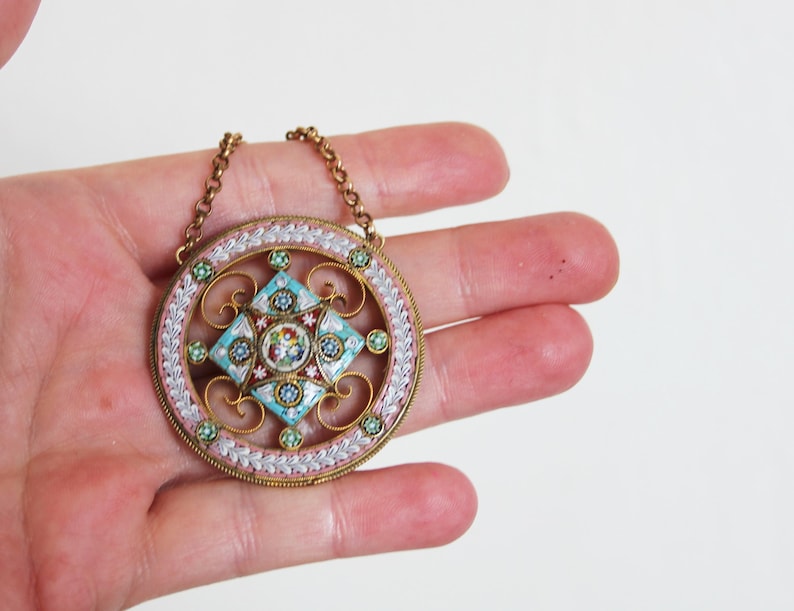 May include: A round pendant necklace with a delicate gold chain. The pendant is made of white, pink, and blue mosaic tiles arranged in a geometric pattern. The pendant is surrounded by a gold filigree design.