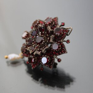 Op de afbeelding: Een zilveren broche met rode granaten in de vorm van een bloem met een parel hanger.