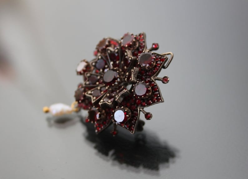 Op de afbeelding: Een zilveren broche met rode granaten in de vorm van een bloem met een parel hanger.