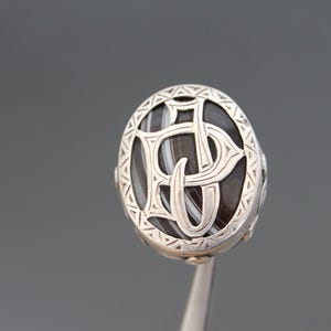 Op de afbeelding: Een zilveren manchetknoop met agaat met een ingewikkeld ontwerp met een monogram. De agaat is een donkerbruine en wit gebande steen.