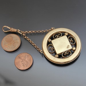 May include: A gold-toned pocket watch fob with a decorative filigree design. The fob has a chain attached to it and a square-shaped locket in the center.