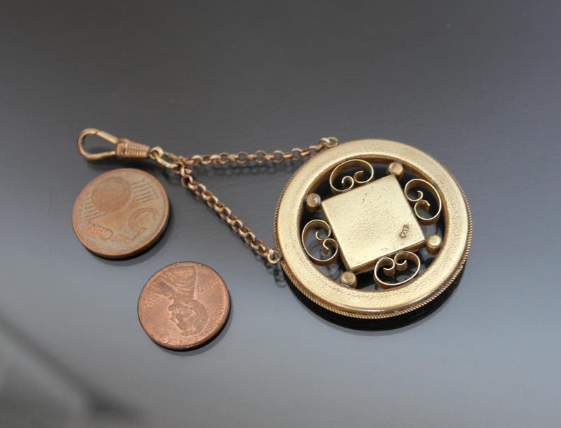 May include: A gold-toned pocket watch fob with a decorative filigree design. The fob has a chain attached to it and a square-shaped locket in the center.