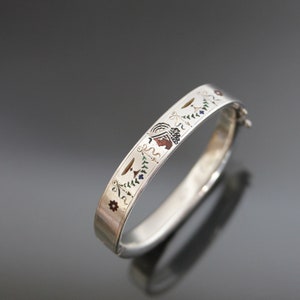 Puede incluir: Un brazalete de plata con un diseño de esmalte colorido que presenta figuras estilizadas y motivos florales.