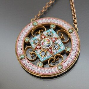 May include: A round pendant necklace with a floral mosaic design in shades of blue, pink, and white. The pendant is surrounded by a delicate gold frame and hangs from a gold chain.