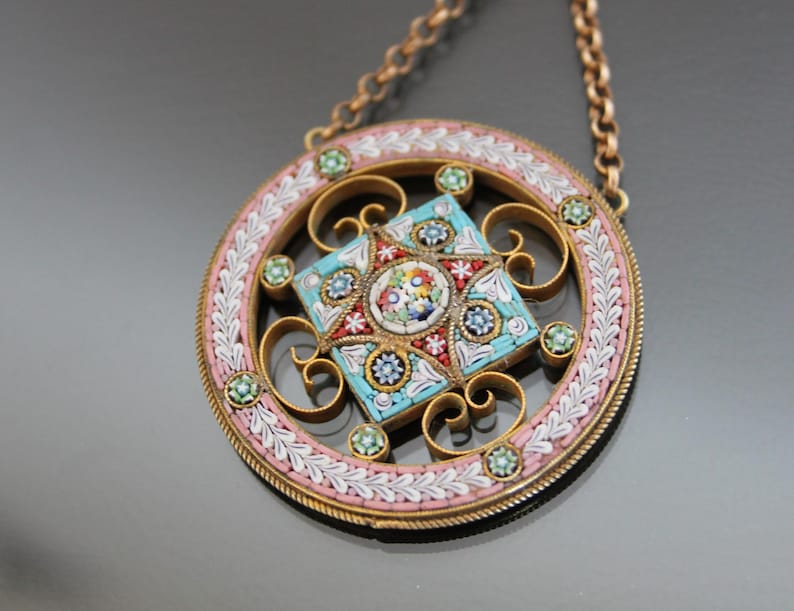 May include: A round pendant necklace with a floral mosaic design in shades of blue, pink, and white. The pendant is surrounded by a delicate gold frame and hangs from a gold chain.