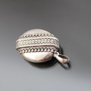 K&ouml;nnte beinhalten: Silbernes ovales Medaillon mit einem filigranen Design am Rand. Das Medaillon hat eine Schlaufe f&uuml;r eine Kette.