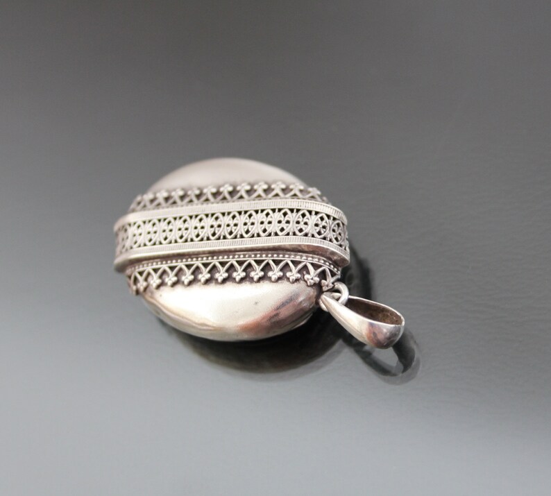 K&ouml;nnte beinhalten: Silbernes ovales Medaillon mit einem filigranen Design am Rand. Das Medaillon hat eine Schlaufe f&uuml;r eine Kette.