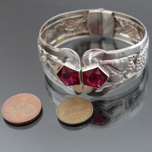 Puede incluir: Un brazalete de plata con un dise&ntilde;o intrincado de uvas y hojas. El brazalete tiene dos acentos de piedras preciosas rojas.