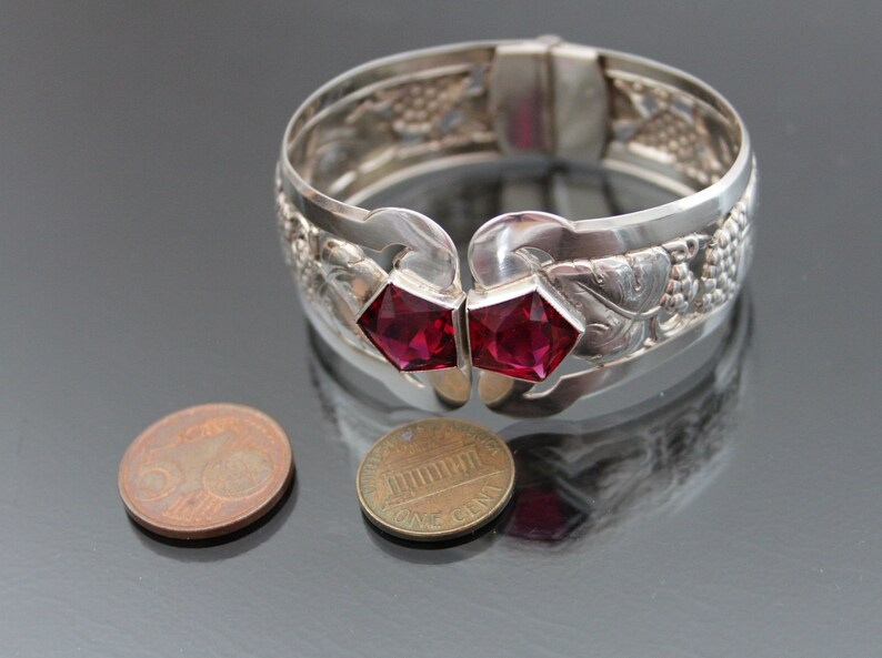 Puede incluir: Un brazalete de plata con un dise&ntilde;o intrincado de uvas y hojas. El brazalete tiene dos acentos de piedras preciosas rojas.