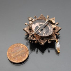 Op de afbeelding: Een gouden broche met een stervormig ontwerp en een parel hanger. De broche heeft een glazen cabochon in het midden met een zwart-wit afbeelding.