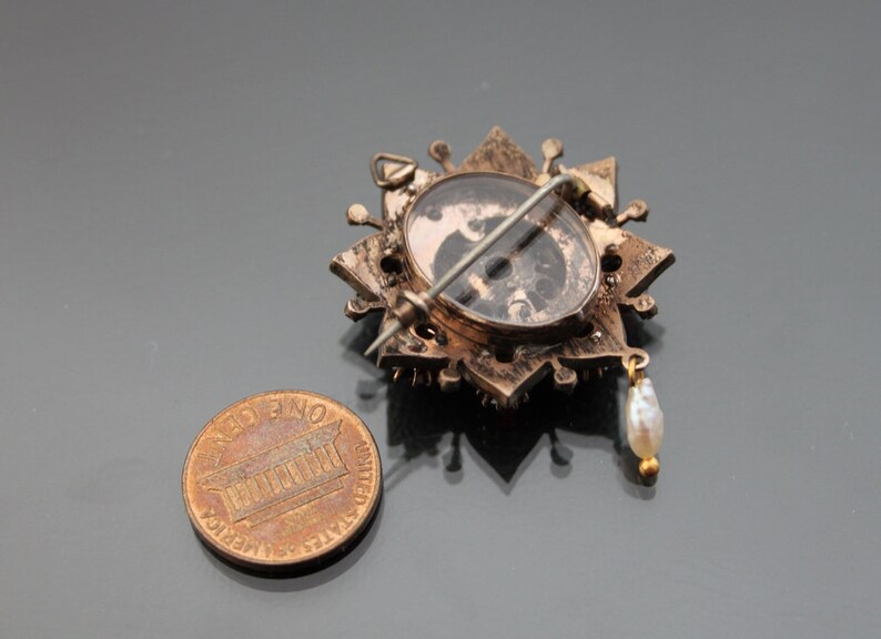 Op de afbeelding: Een gouden broche met een stervormig ontwerp en een parel hanger. De broche heeft een glazen cabochon in het midden met een zwart-wit afbeelding.