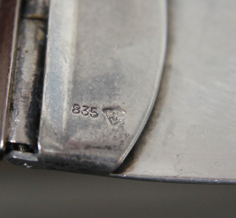 May include: Close-up of a silver metal object with the number "835" and a small triangle symbol stamped on it.