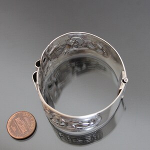May include: A silver bracelet with an intricate, openwork design. The bracelet is hinged and has a clasp. The bracelet is about the size of a US penny.