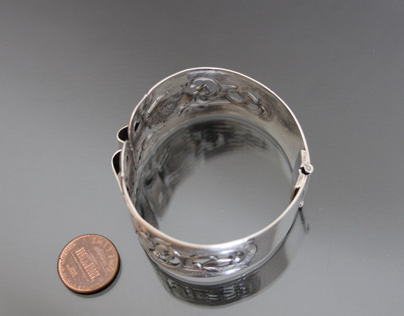 May include: A silver bracelet with an intricate, openwork design. The bracelet is hinged and has a clasp. The bracelet is about the size of a US penny.