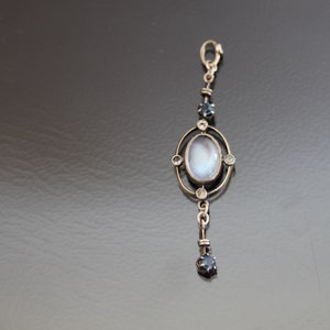 Könnte beinhalten: Ein silberner Anhänger mit einem ovalen Mondstein-Cabochon, der in einen silbernen Rahmen mit vier kleinen Diamanten gefasst ist. Der Anhänger hängt an einer silbernen Kette mit einem kleinen blauen Saphir am unteren Ende.