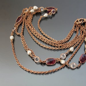 Op de afbeelding: Een meerstrengs ketting met een gouden ketting en rode en heldere edelstenen. De ketting heeft een sluiting met een ronde hanger.