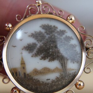 May include: A gold locket with a delicate, ornate design. The locket contains a miniature scene of a church, a tree, and a lake, created with hair art.