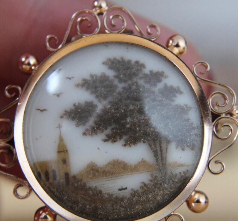 May include: A gold locket with a delicate, ornate design. The locket contains a miniature scene of a church, a tree, and a lake, created with hair art.