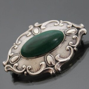 May include: A silver brooch with an oval-shaped green stone set in a decorative frame.